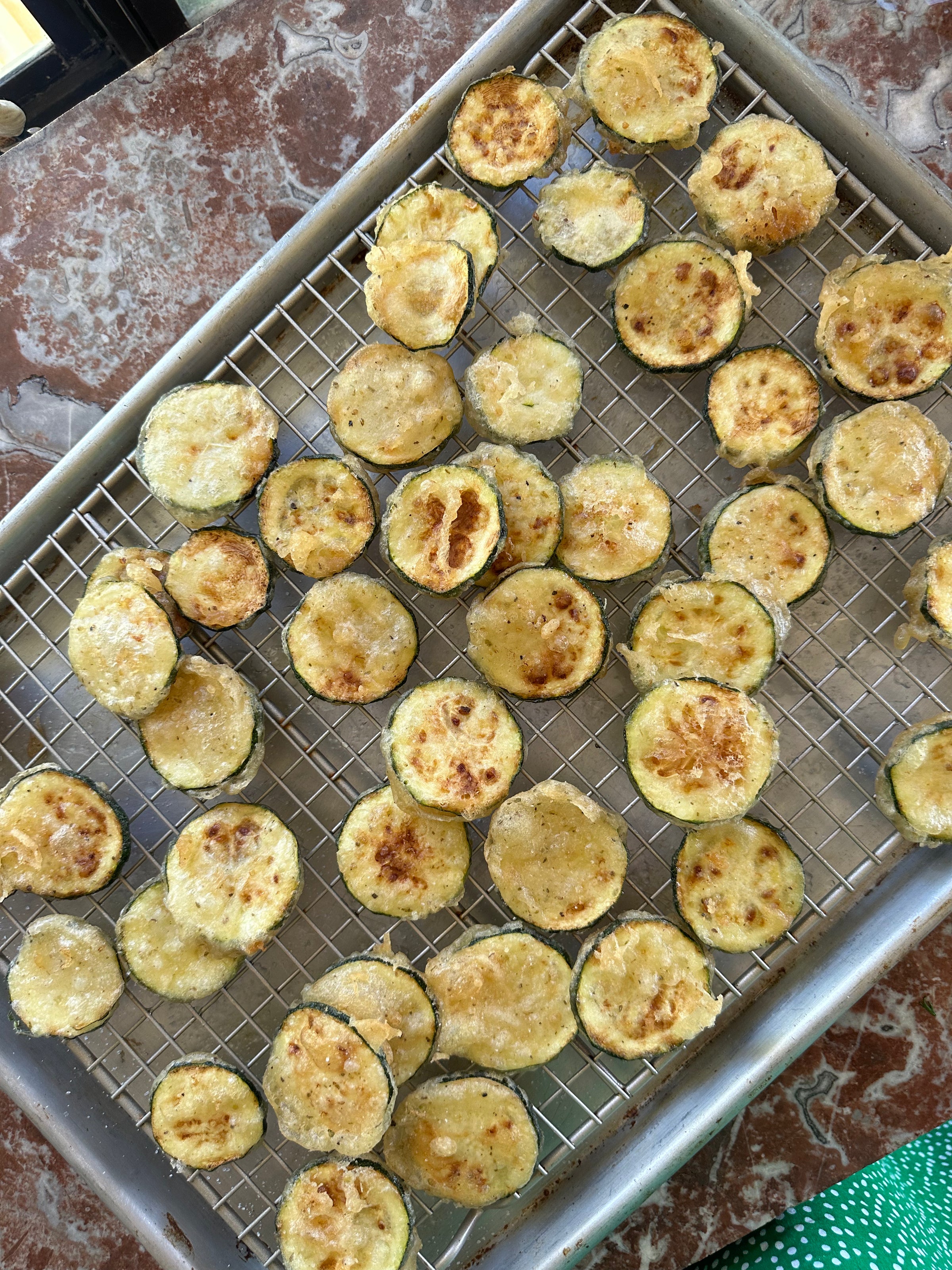 A baking sheet with a wire rack holds many golden-brown, round zucchini slices, arranged in a single layer on a marbled countertop. The zucchini appear to be roasted or baked.