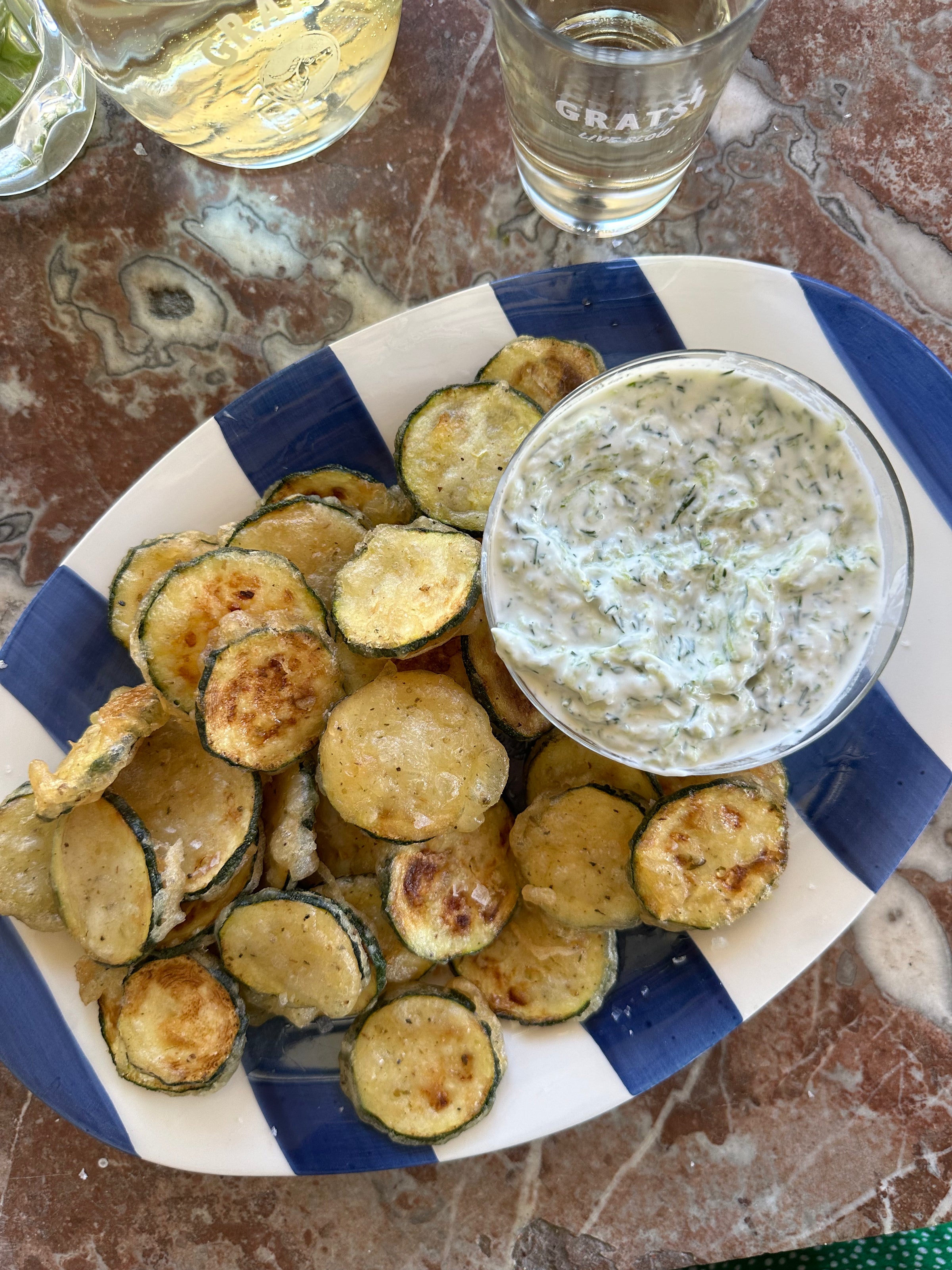 A plate of crispy fried zucchini slices is served with a bowl of creamy herb dip on a blue and white dish, set on a marbled table next to a glass of clear liquid.