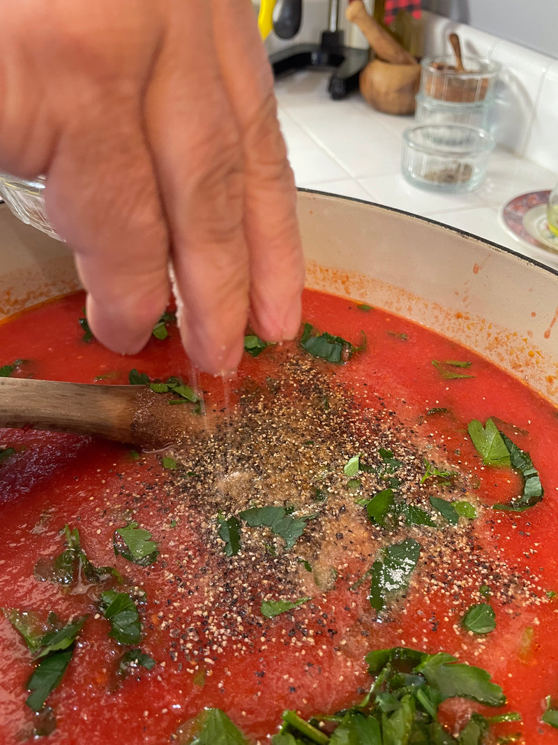A hand sprinkles salt into a pot of tomato sauce with chopped herbs and ground pepper, while a wooden spoon rests in the red sauce on a kitchen stovetop.