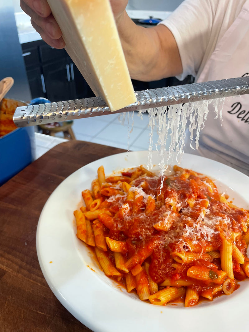 A person grates a block of cheese over a plate of penne pasta topped with tomato sauce, using a metal grater. The pasta is served on a white plate on a wooden table.