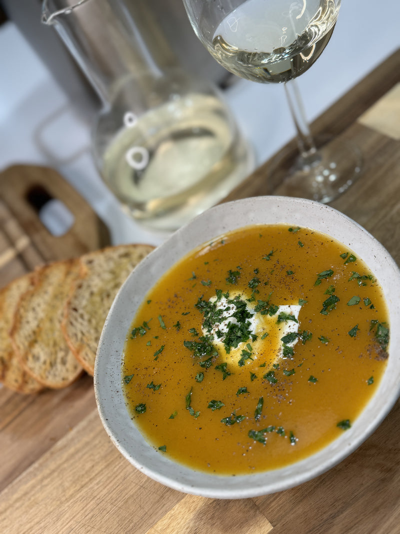 A bowl of orange-colored soup garnished with cream and herbs sits on a wooden board, next to three slices of bread, a glass of white wine, and a glass carafe.