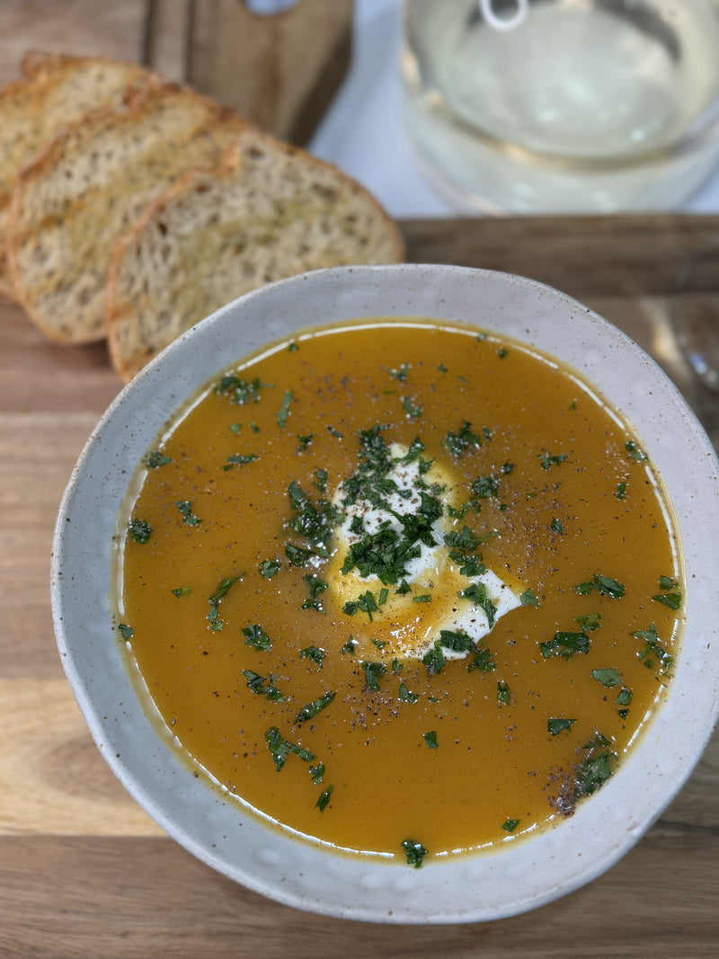 A bowl of creamy orange soup garnished with herbs and a dollop of cream, served on a wooden board with several slices of bread and a glass of white wine in the background.