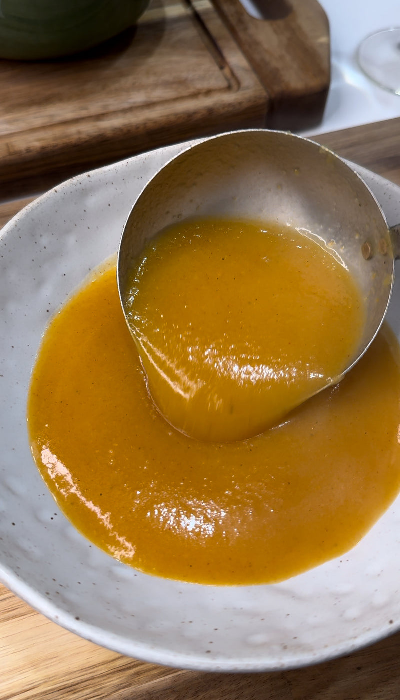 A ladle pours thick, golden-orange soup or sauce into a white bowl on a wooden surface.