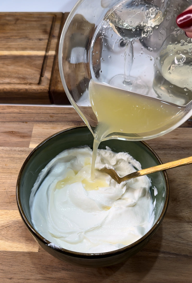 A hand pours lemon juice from a glass measuring cup into a green bowl of thick white yogurt on a wooden surface, mixing with a gold spoon.