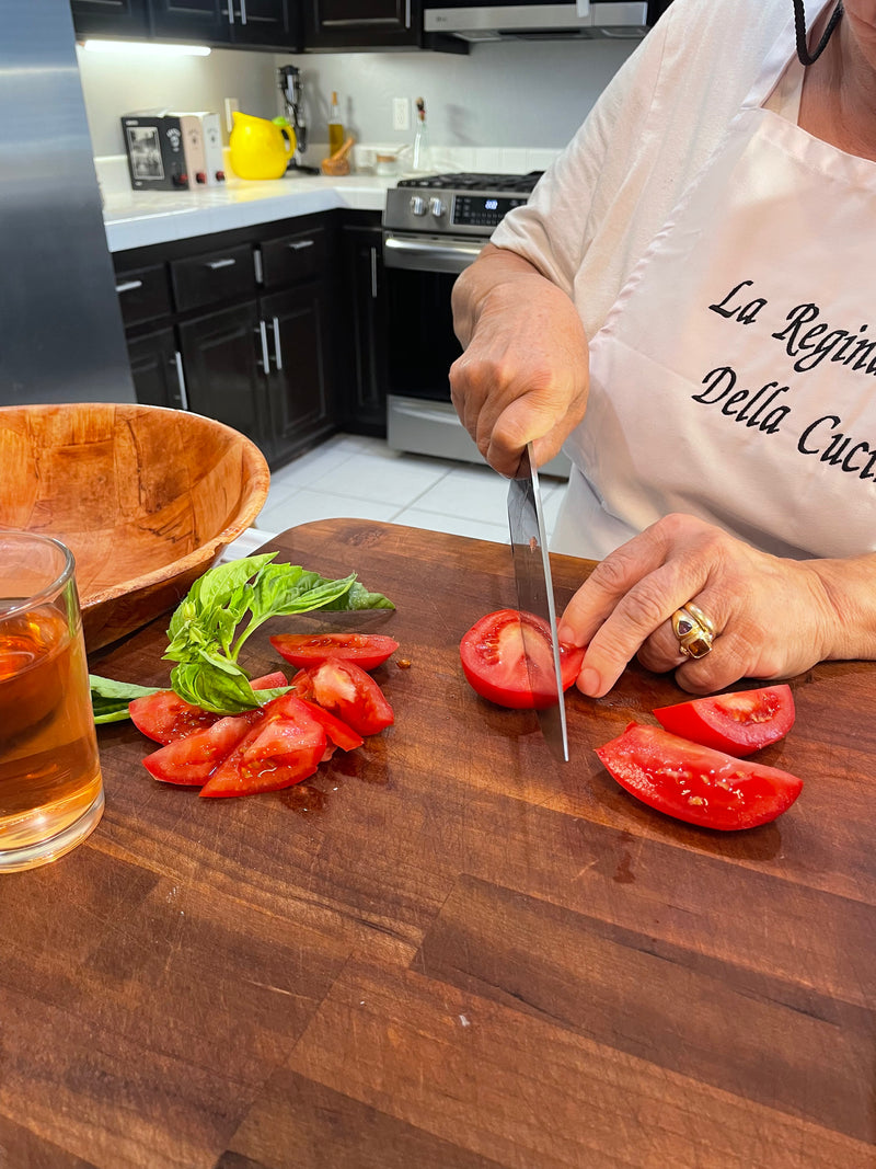 A person wearing a white apron cuts ripe tomatoes on a wooden board in a kitchen. Fresh basil, a wooden bowl, and a glass of tea are nearby. Only the person’s hands and torso are visible.