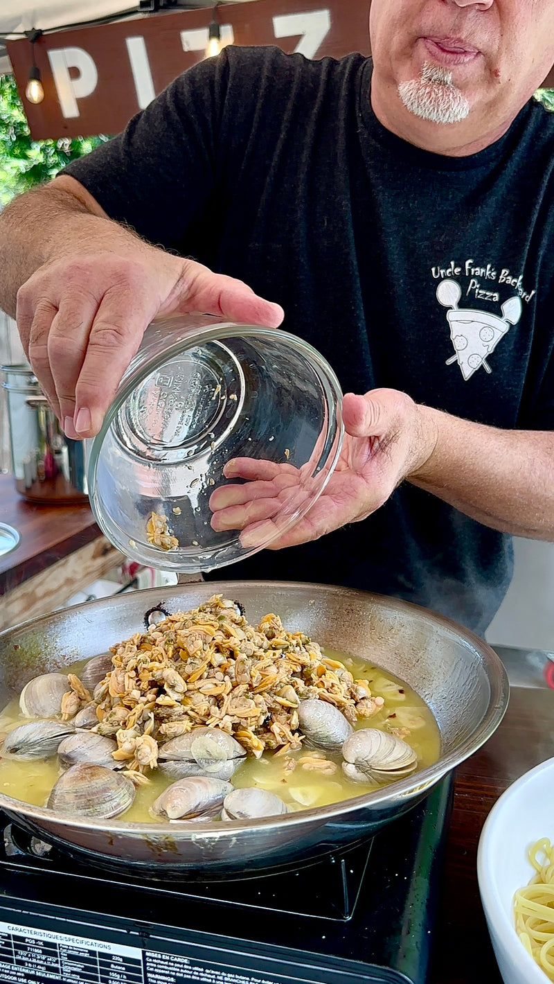 A person pours chopped clams from a glass bowl into a skillet filled with clams and broth. The person wears a black shirt with a pizza logo that says Uncle Franks Backyard Pizza.
