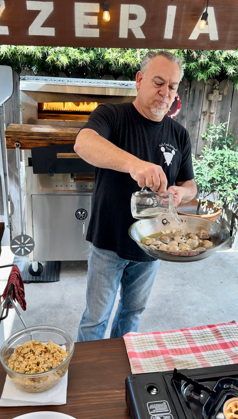 A man pours liquid from a glass pitcher into a skillet of clams outdoors, standing beside a wood-fired pizza oven, with ingredients and a red-checked cloth on the table in front of him.