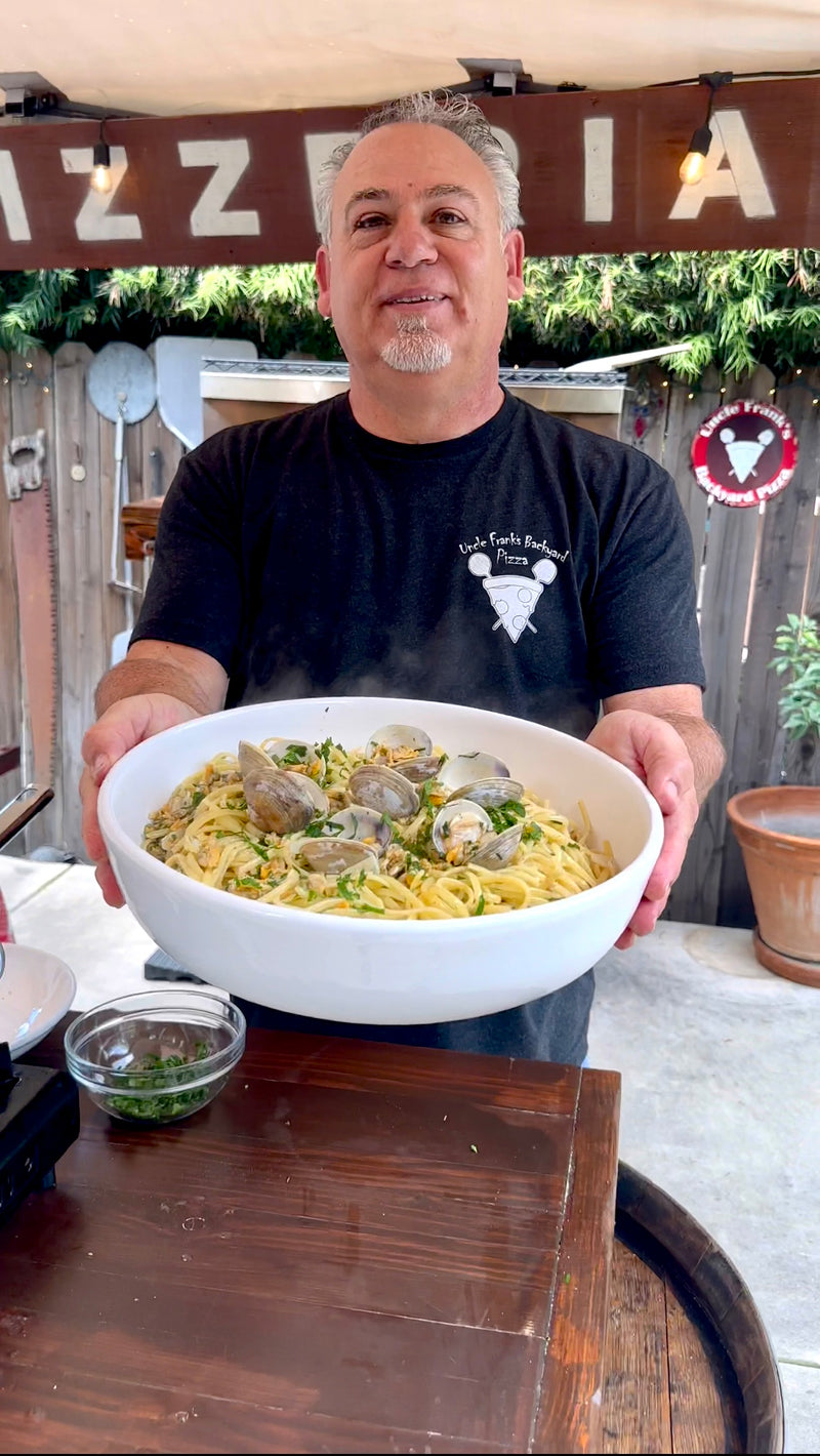 A man in a black T-shirt stands outdoors, smiling and holding a large white bowl filled with pasta and clams. Potted plants and wooden fencing are in the background. A small bowl with herbs sits on the table in front of him.