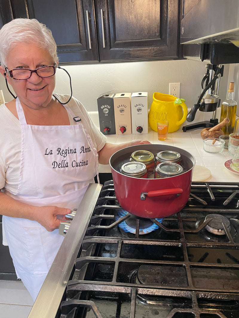 An older woman wearing glasses and a white apron stands by a stove, smiling. Three glass jars are boiling in a red pot on the burner. The kitchen has dark cabinets and a yellow lemonade pitcher on the counter.