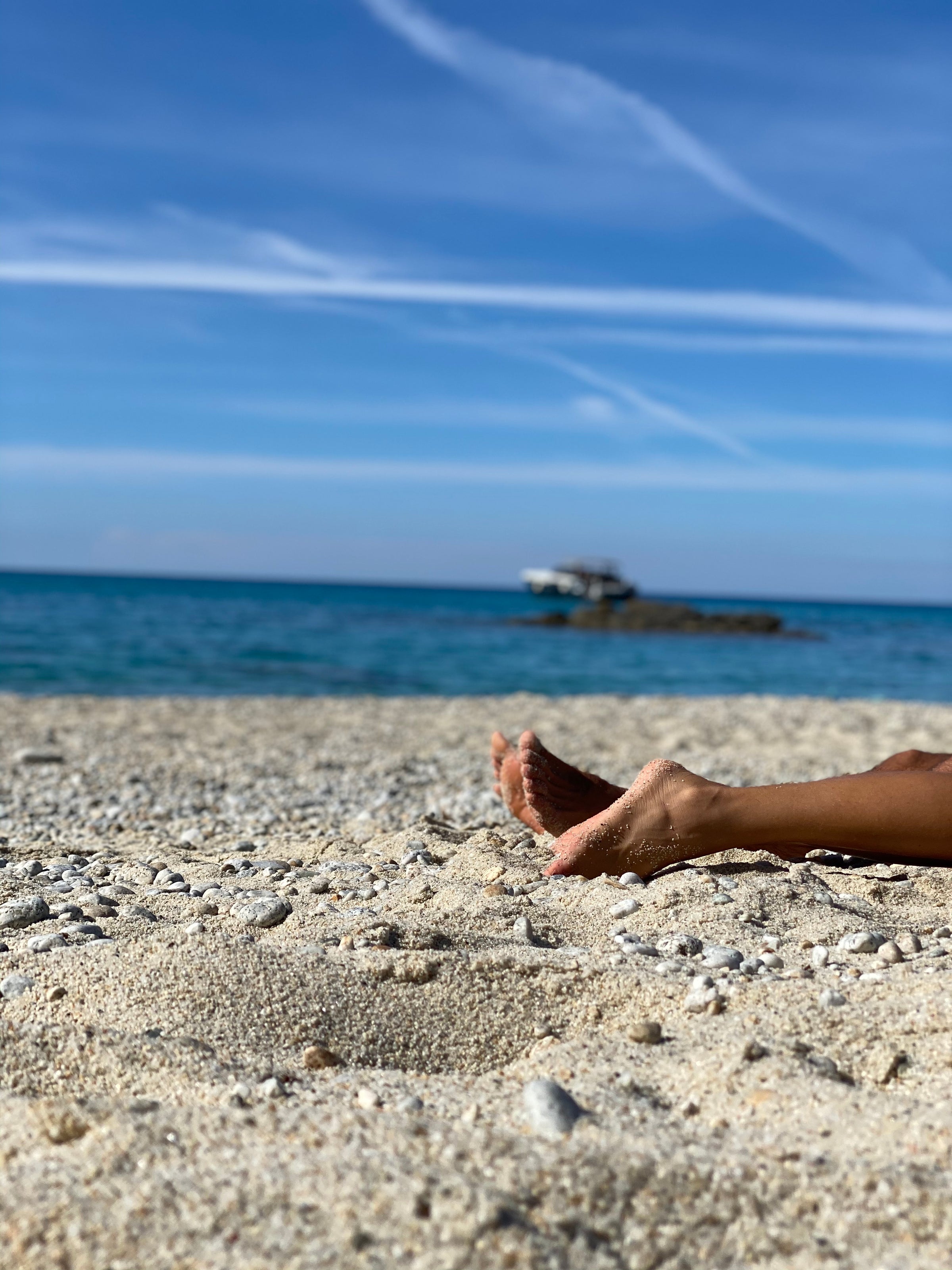 Bare feet partially buried in sandy beach in the foreground, with a calm blue ocean and a boat floating in the distance under a clear sky with wispy clouds.