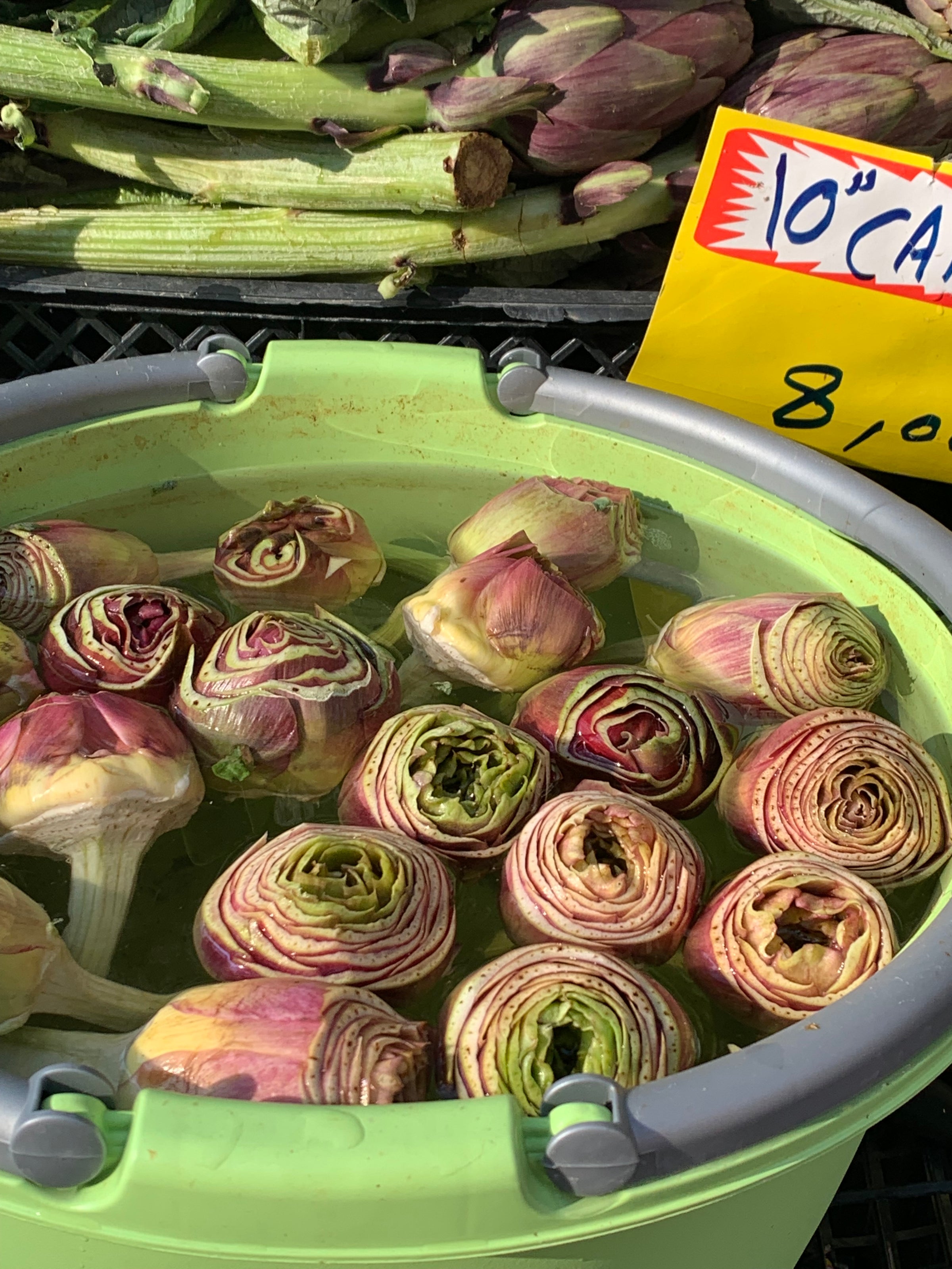 A green bucket filled with trimmed artichokes soaked in water, displayed at an outdoor market. Whole artichokes and a yellow price sign are visible in the background.