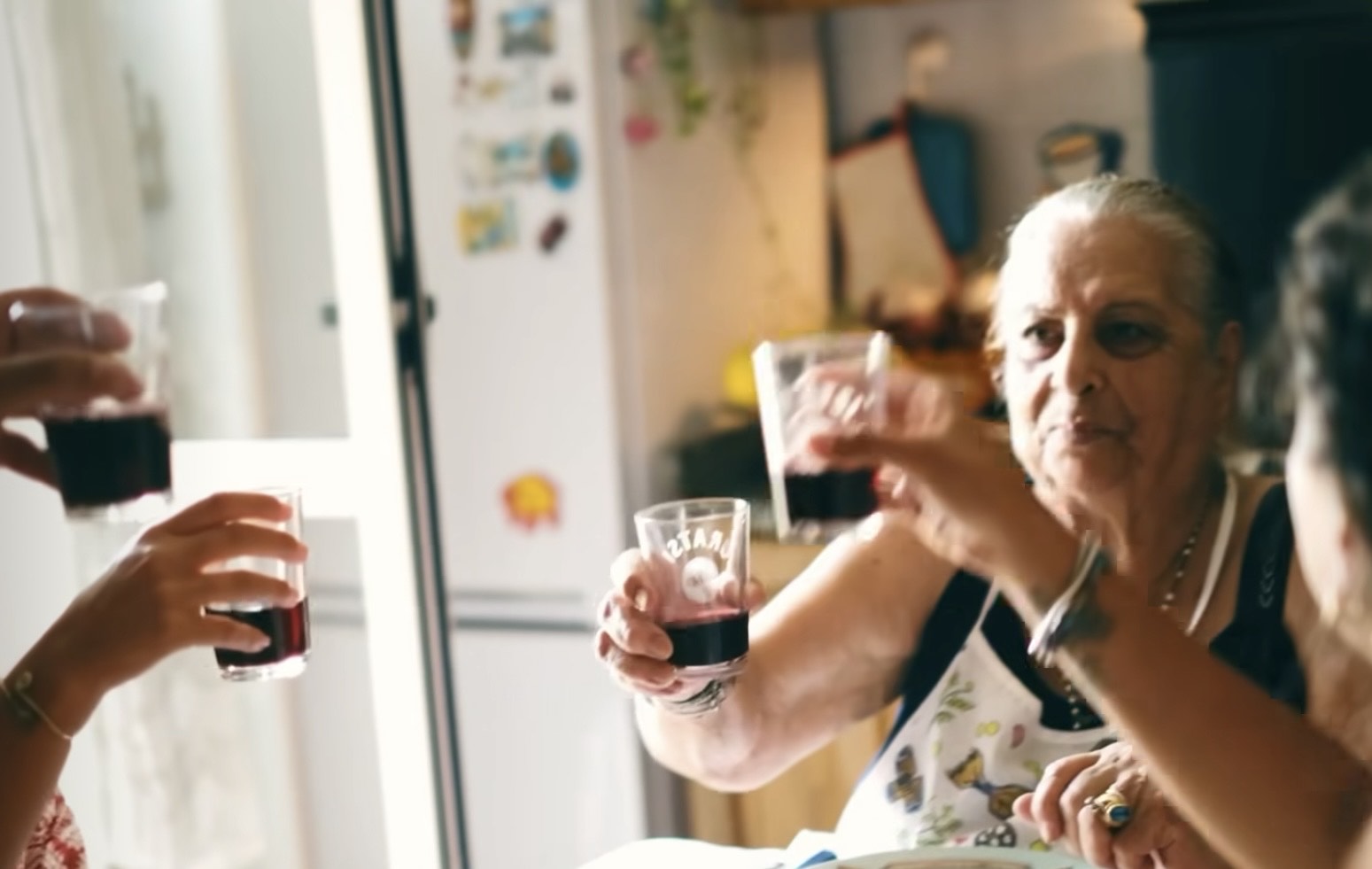 Three people raise glasses of red wine at a table in a cozy kitchen. An older woman wearing an apron smiles warmly, joining the toast. The background includes a refrigerator with magnets and soft, natural light.