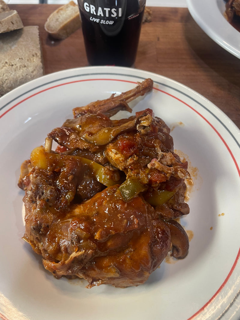 A plate of braised meat with vegetables in a rich sauce, served on a white plate with a red rim. Slices of bread and a dark drink in a glass labeled GRATSI are visible in the background.
