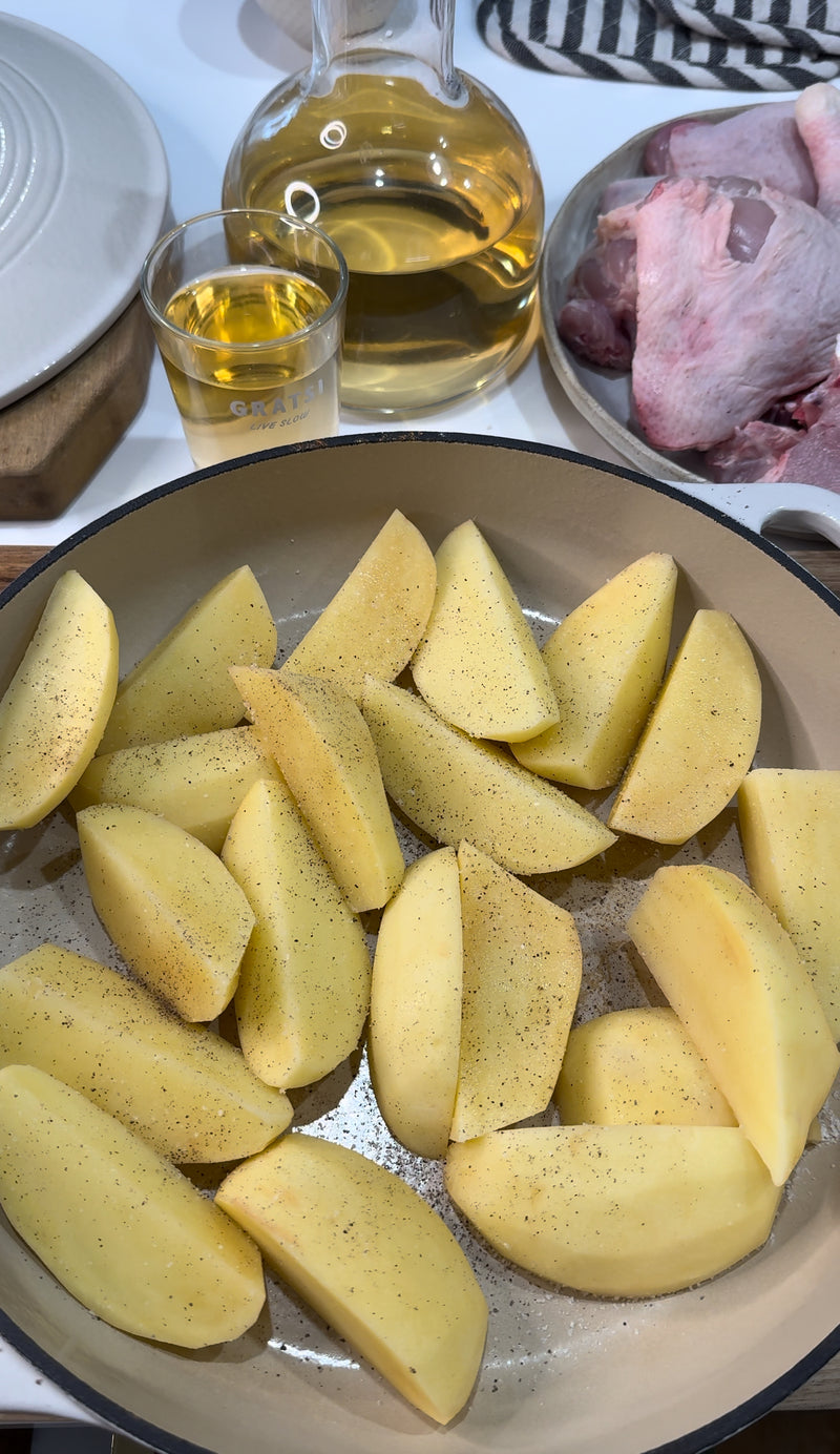 Sliced potatoes seasoned with pepper are arranged in a pan, with a glass and carafe of white wine and raw poultry in the background, ready for cooking.