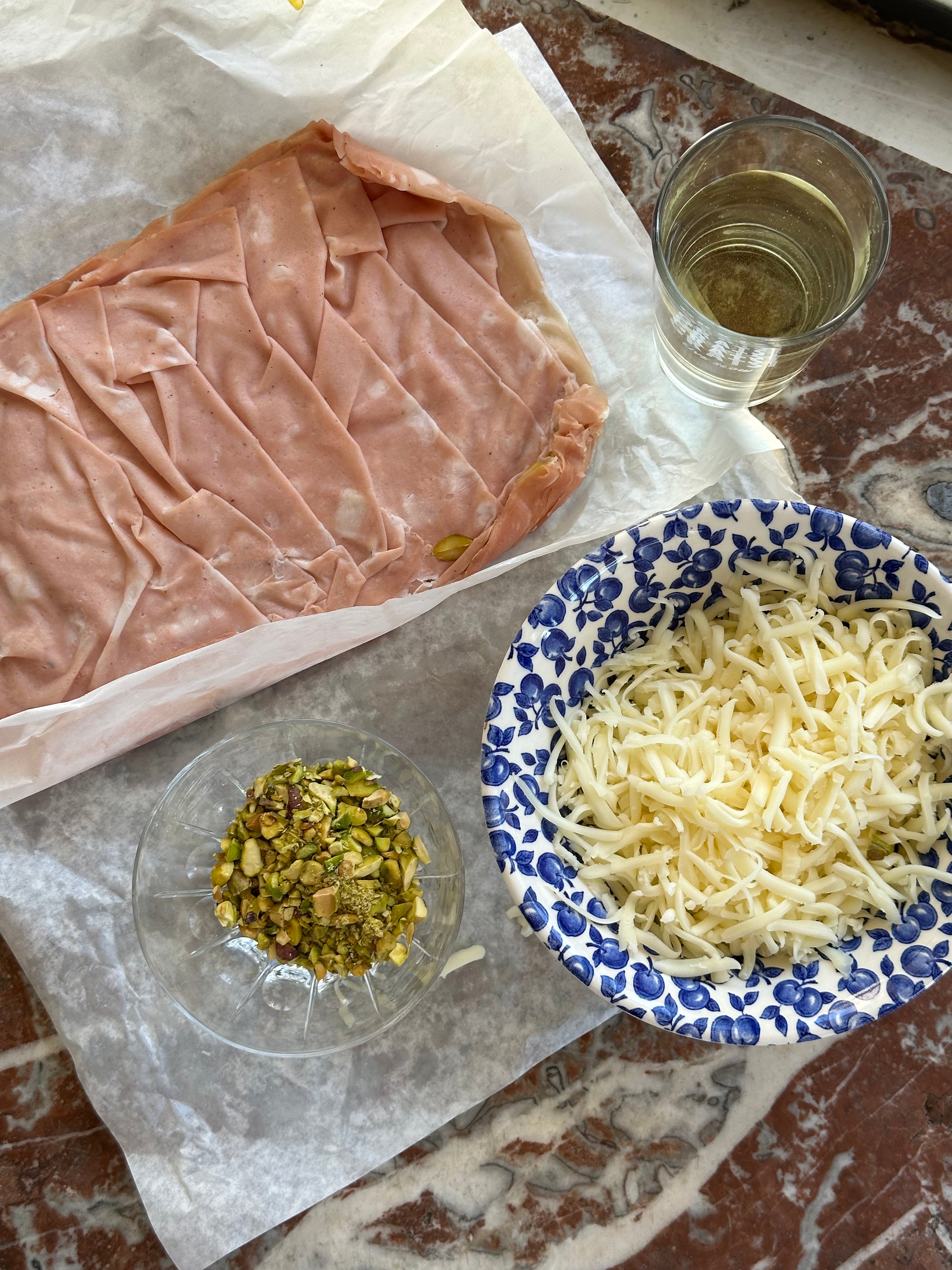 A block of mortadella on parchment, a glass of white wine, a bowl of shredded cheese, and a small dish of chopped pistachios are arranged on a marbled brown countertop.