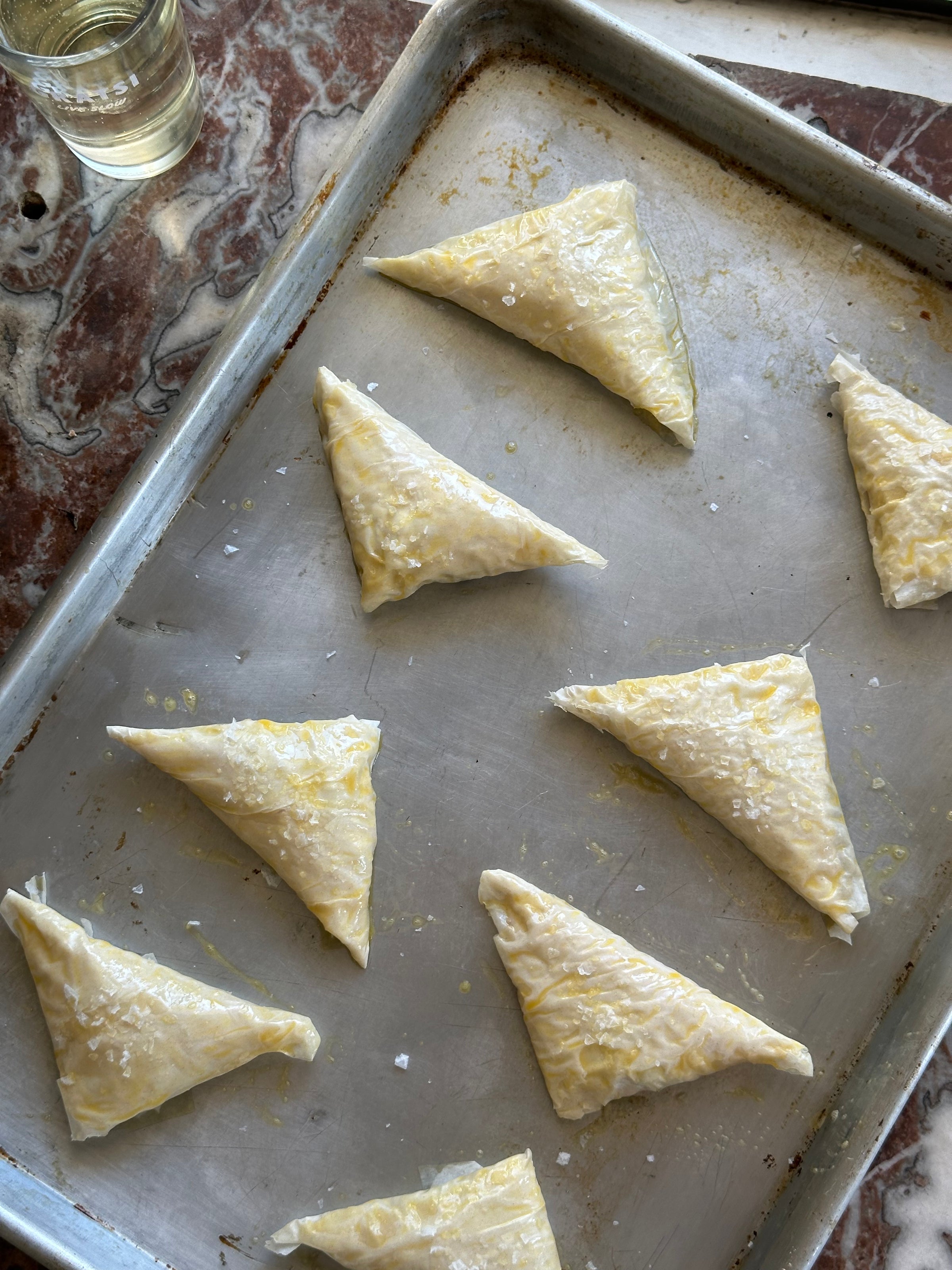 Eight unbaked, triangular phyllo dough pastries are arranged on a metal baking sheet, brushed with egg wash and sprinkled with salt. A glass of liquid sits on the marbled countertop beside the tray.