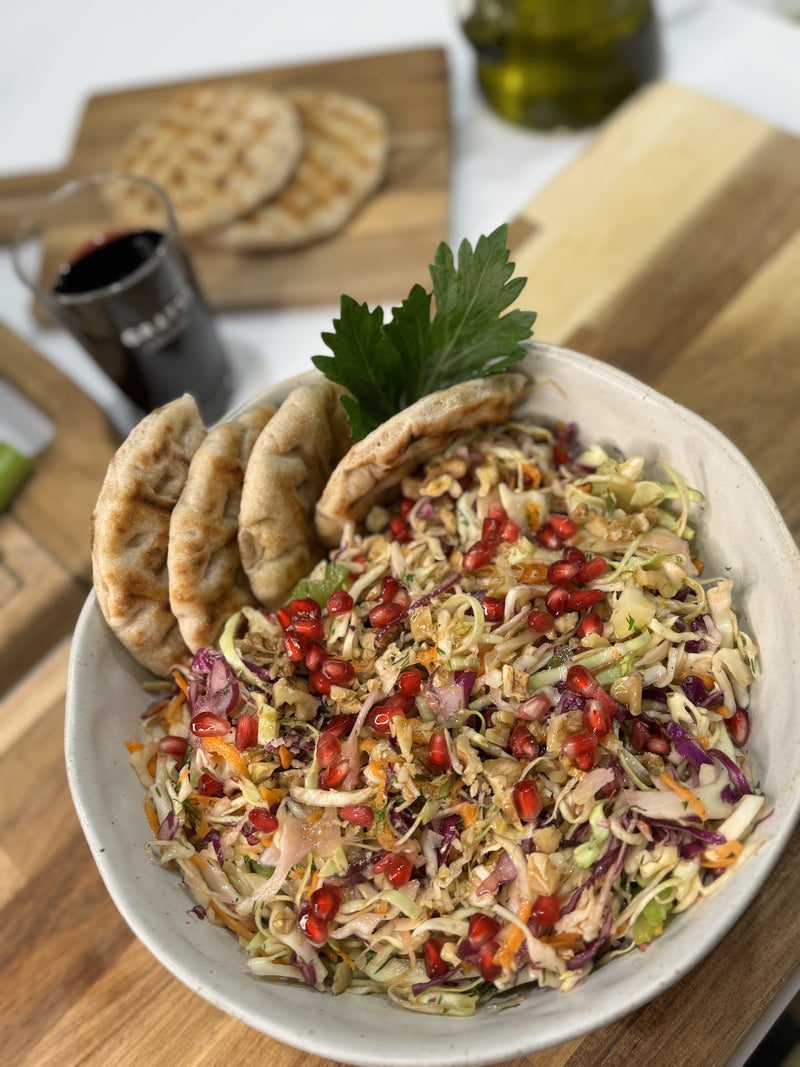A bowl of colorful shredded cabbage salad topped with pomegranate seeds and herbs, garnished with pita bread slices. In the background, there’s a glass of red drink, olive oil, and more pita on a wooden board.