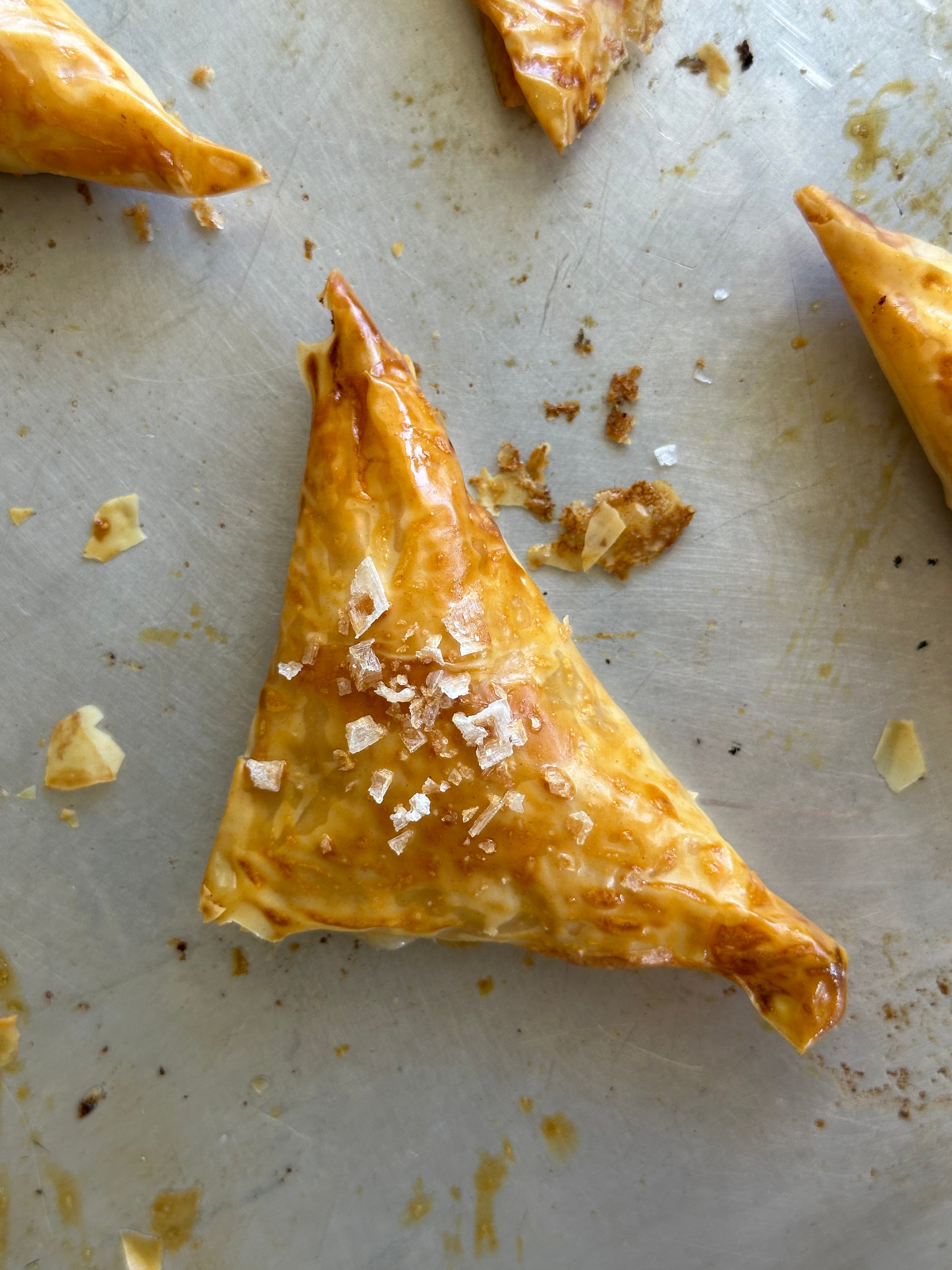 A golden-brown, triangular pastry topped with flaky sea salt, resting on a metal baking sheet with scattered crumbs.