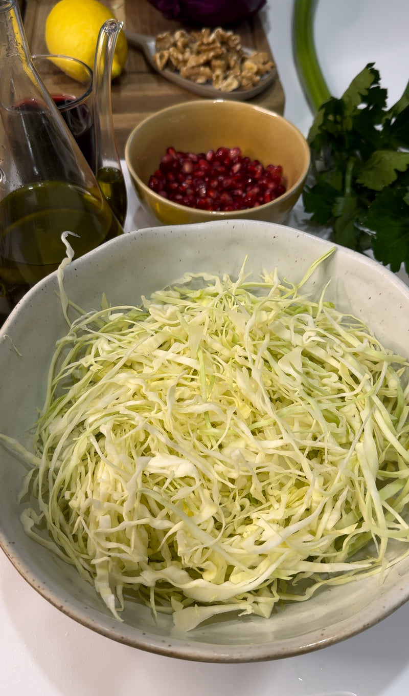 A bowl of finely shredded cabbage is in the foreground, with a bottle of olive oil, a bowl of pomegranate seeds, fresh parsley, and walnuts visible in the background on a kitchen counter.