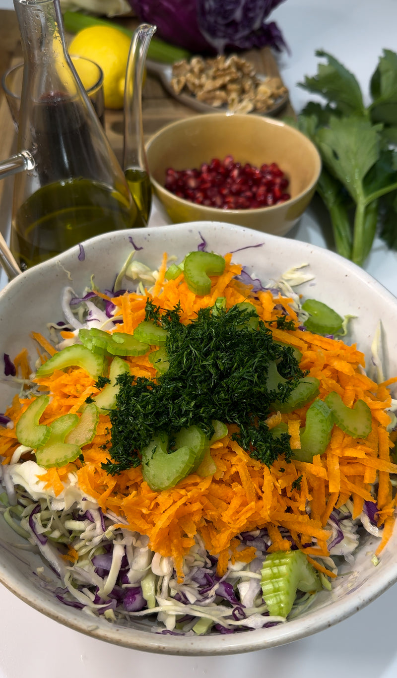 A bowl of fresh salad with shredded cabbage, grated carrots, celery slices, and chopped herbs, surrounded by olive oil, pomegranate seeds, walnuts, a lemon, and celery leaves on a table.