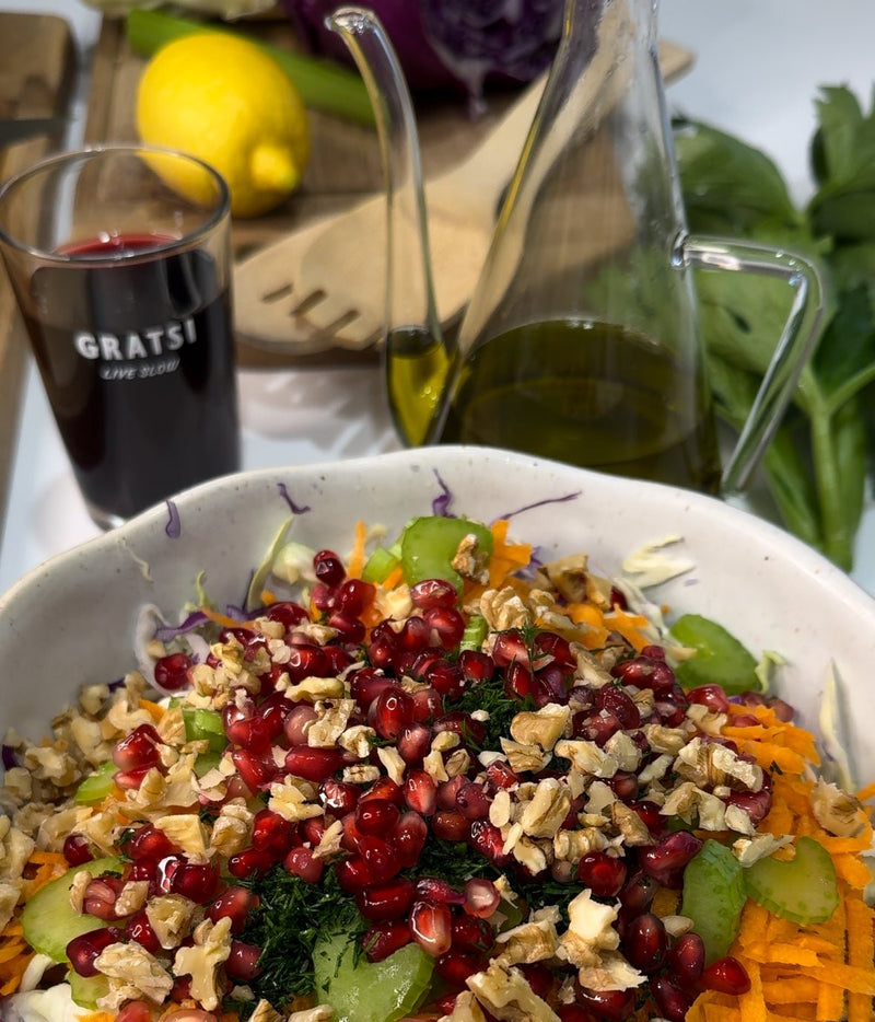 A salad topped with pomegranate seeds, walnuts, cucumber, and herbs sits in a white bowl. Nearby are a glass of red wine, a bottle of olive oil, a lemon, and fresh green leaves on a wooden board.