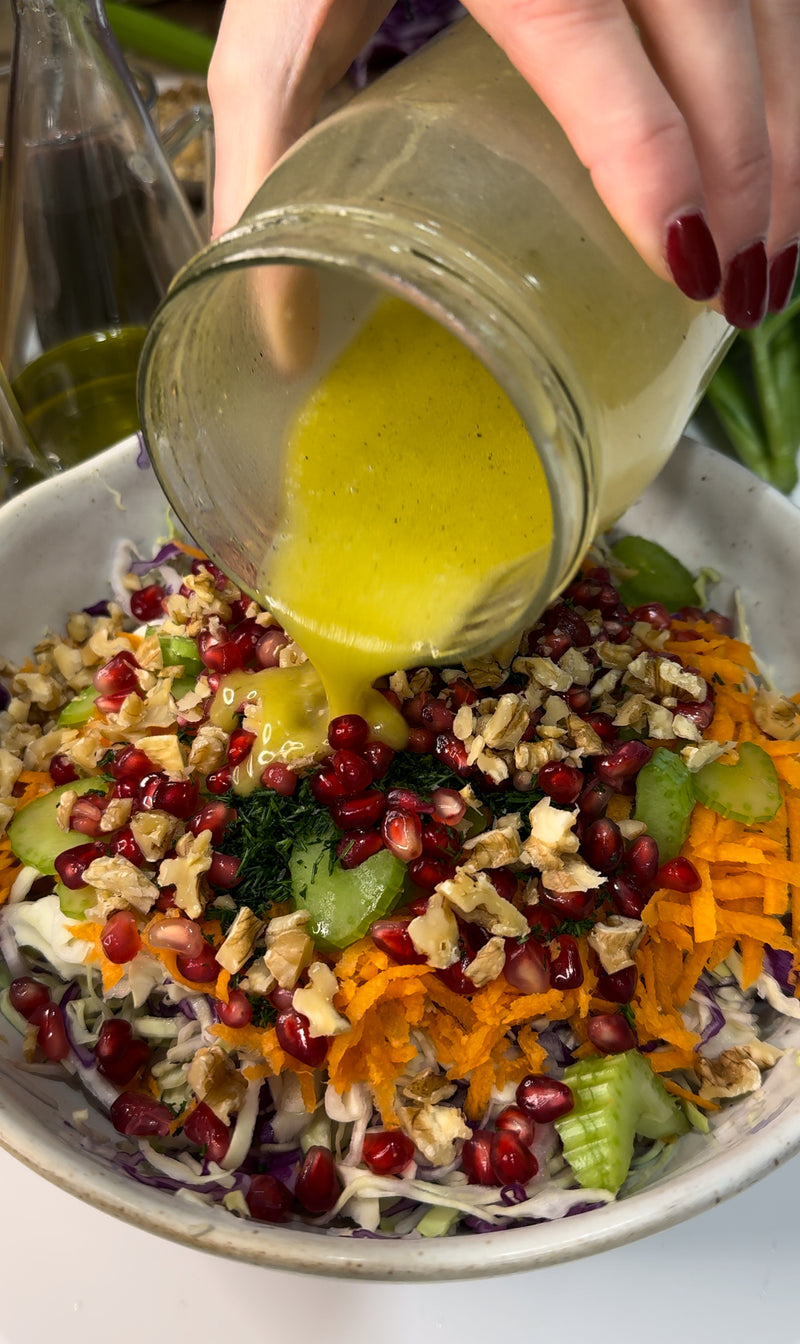 A hand pours yellow salad dressing from a jar onto a colorful salad with shredded carrots, pomegranate seeds, chopped walnuts, celery, and greens in a white bowl.