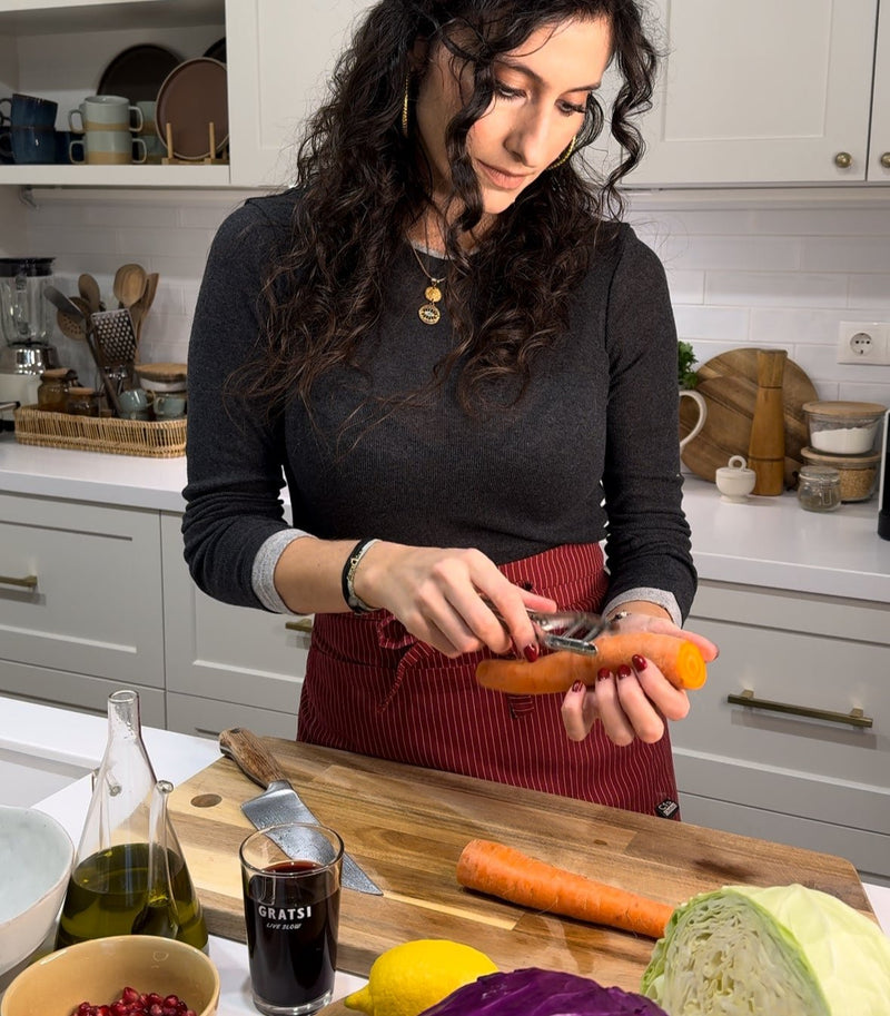 A woman in a kitchen peels a carrot over a wooden cutting board, surrounded by fresh vegetables, a glass of red wine, and kitchen utensils. She wears a dark long-sleeve top and a red apron.