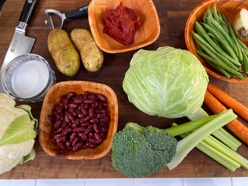 A wooden table with various vegetables and ingredients: cabbage, broccoli, celery, carrots, green beans, cauliflower, potatoes, an onion, red kidney beans, tomato paste, a bowl of salt, a meat cleaver, and a vegetable peeler.