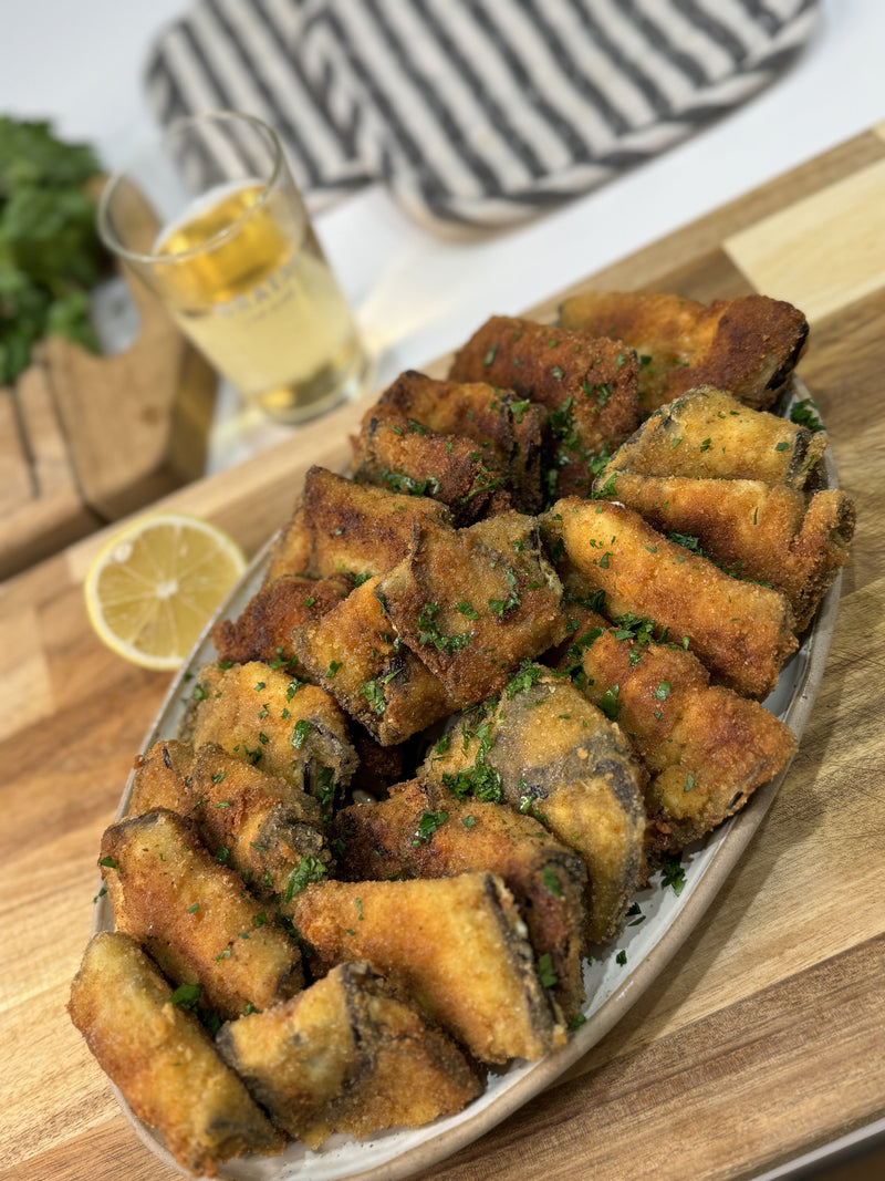 A plate of golden-brown, breaded and fried fish pieces garnished with chopped herbs, set on a wooden board. A lemon slice and a glass of sparkling drink are in the background.