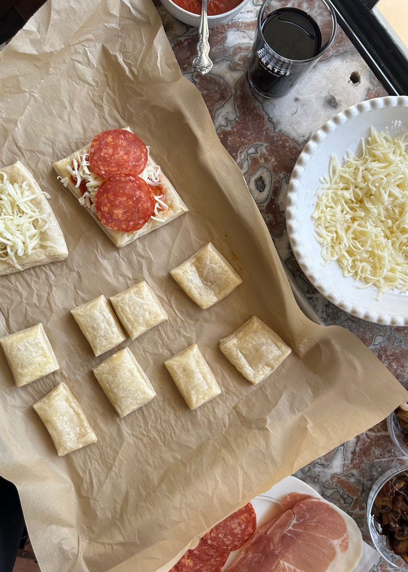 A parchment-lined baking sheet with unbaked dough squares and two pizza bread pieces topped with cheese and pepperoni, surrounded by bowls of shredded cheese, sauce, and a glass of red drink on a marble surface.