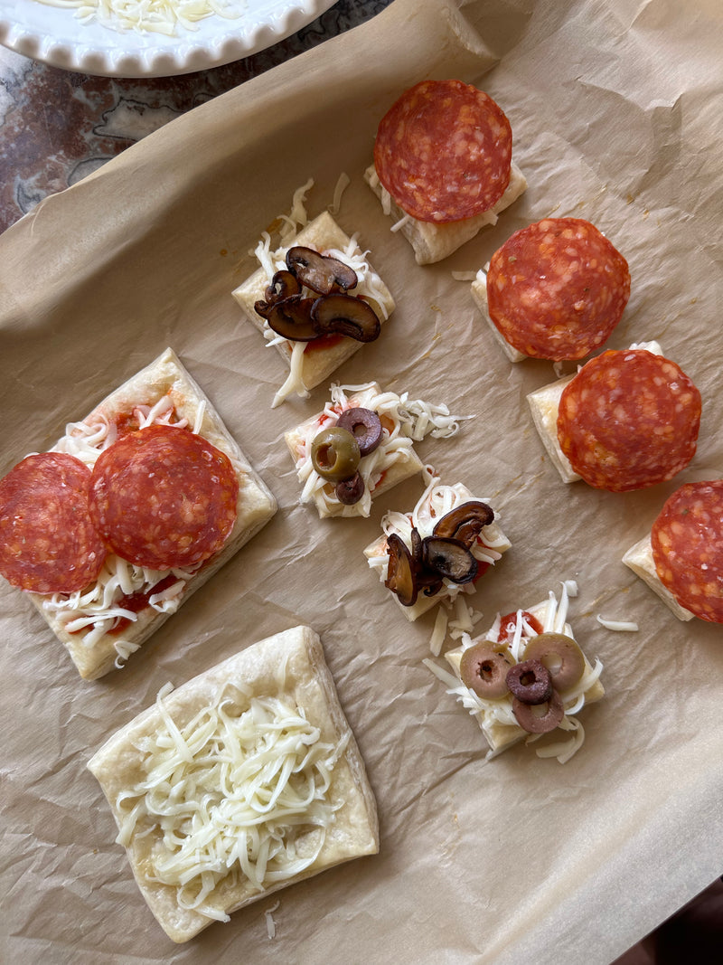 Several pieces of square, uncooked dough on parchment paper, topped with mozzarella cheese, pepperoni slices, mushrooms, and olives, arranged in preparation for baking.