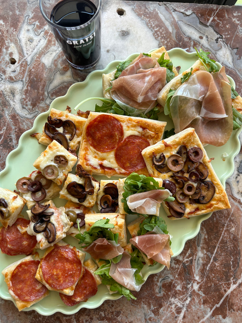 A tray with square slices of pizza topped with pepperoni, mushrooms, and olives, alongside sandwiches with prosciutto and greens. A glass of dark soda is nearby, all on a marbled table.