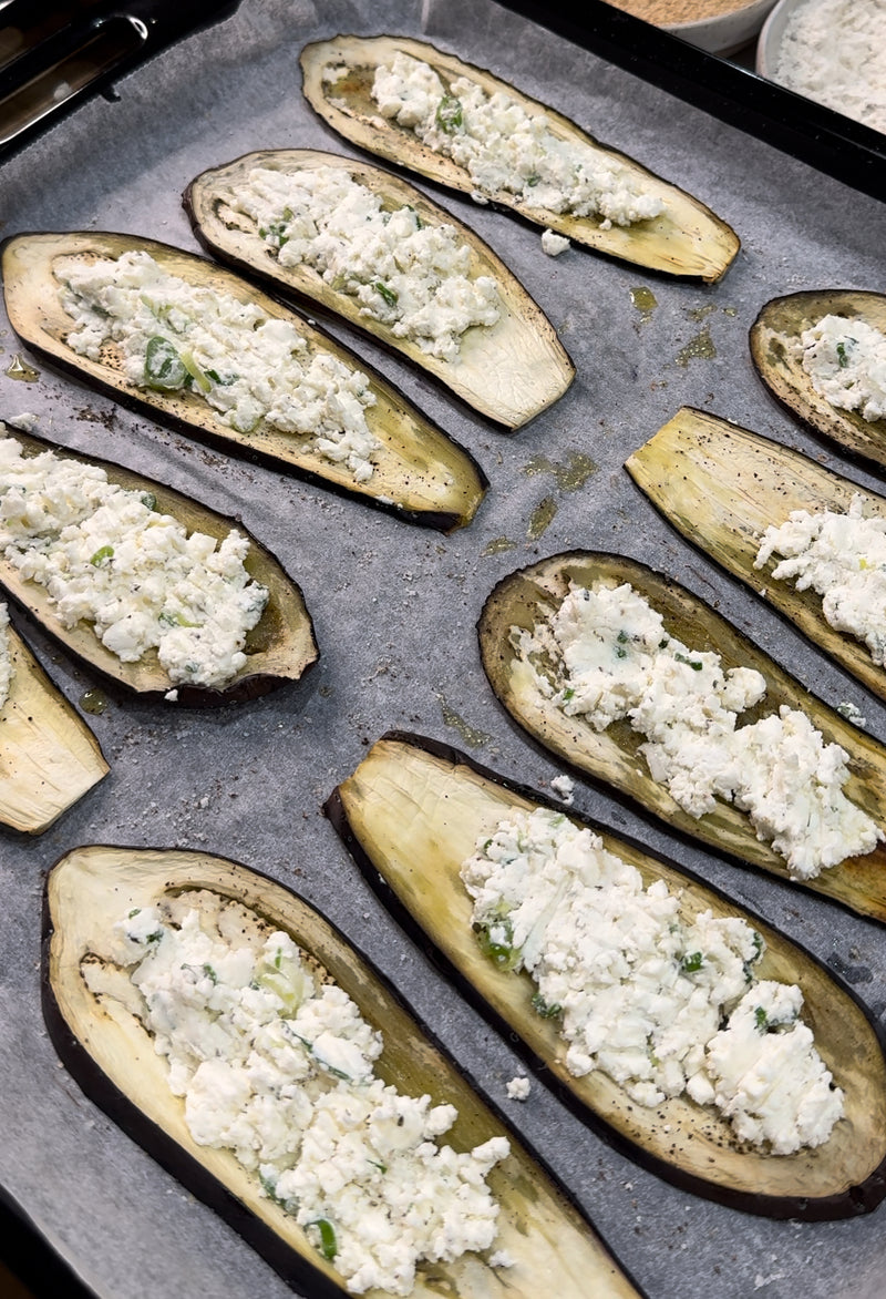 Sliced eggplant halves on a baking tray lined with parchment paper, each topped with a white ricotta cheese and herb mixture, ready to be baked.