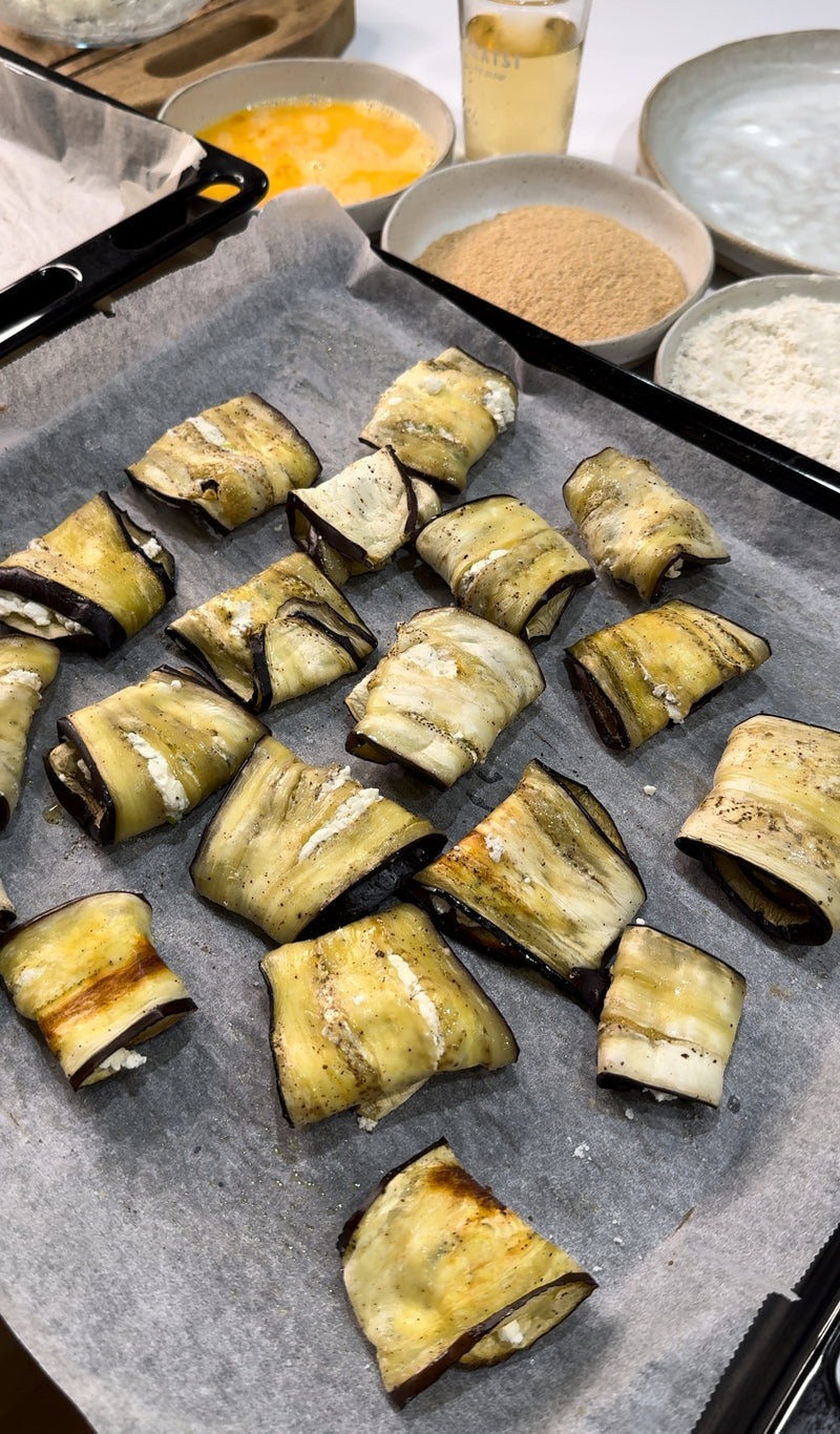 Slices of eggplant rolled around a filling are arranged on a parchment-lined baking tray. In the background are bowls with flour, breadcrumbs, and beaten eggs, suggesting preparation for baking or frying.