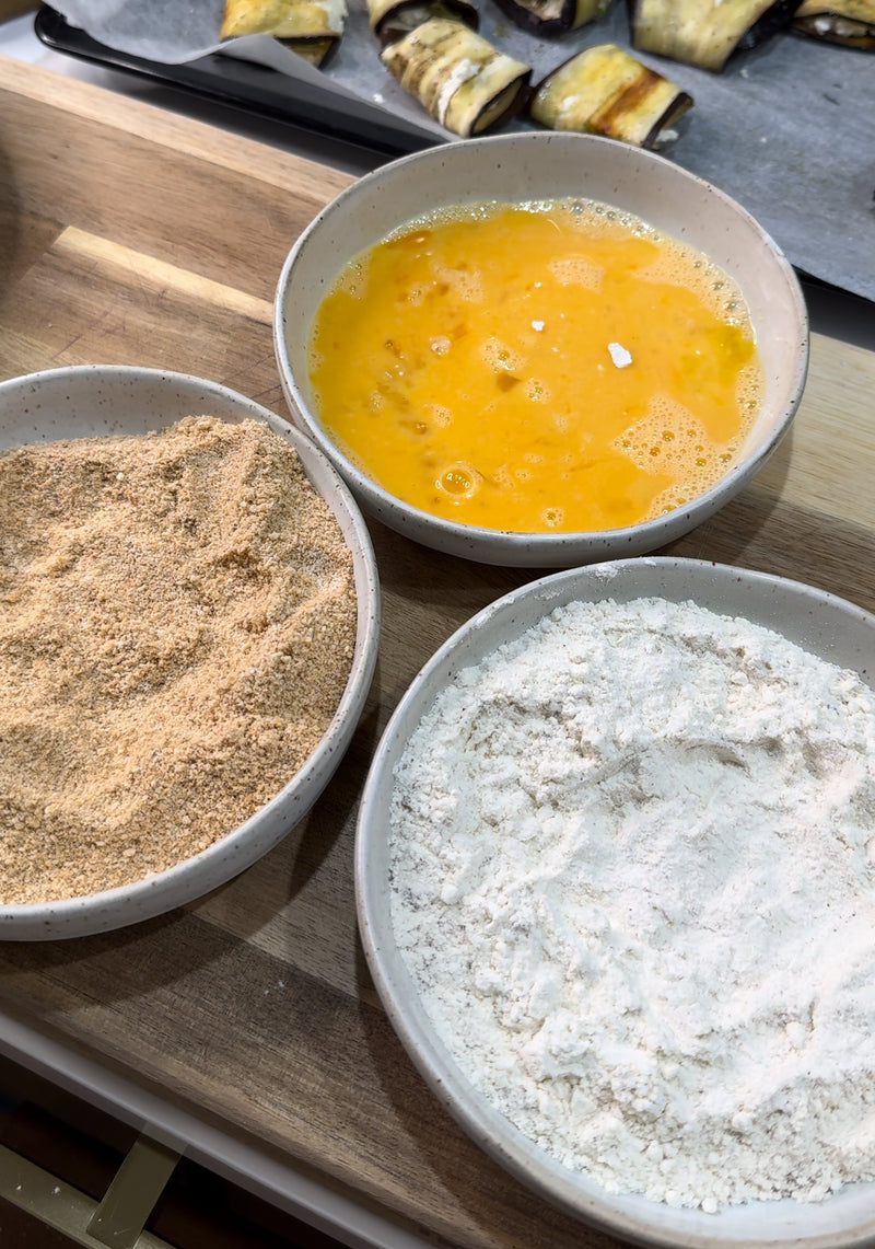 Three bowls on a wooden surface: one with flour, one with beaten eggs, and one with breadcrumbs, ready for coating food items.