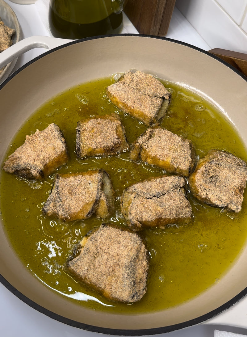 Several pieces of breaded fish are being shallow-fried in hot oil in a large beige pan. The fish pieces are golden brown and partially submerged in oil. A bottle of olive oil is visible in the background.