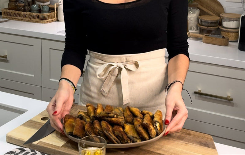 A person wearing a beige apron holds a large plate filled with baked pastries in a kitchen, with white cabinets and various kitchen items in the background.