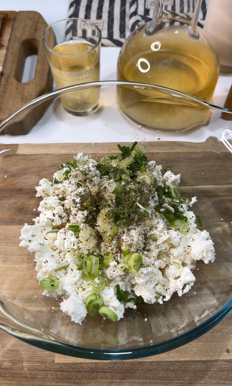 A glass bowl contains crumbled white cheese, chopped green onions, herbs, and black pepper on a wooden surface. Behind it are a glass of white wine, a carafe with liquid, and a striped towel.
