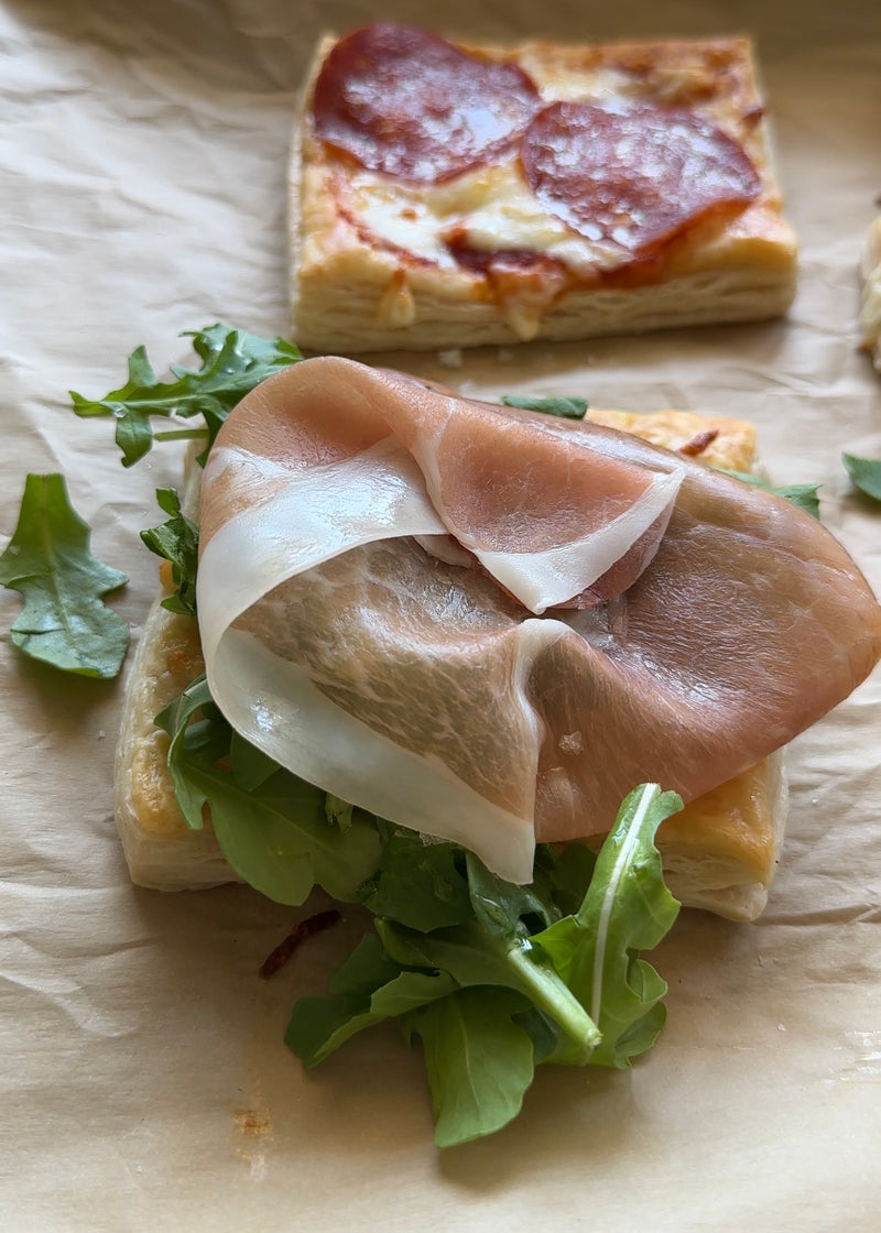 A close-up of a puff pastry square topped with arugula and a slice of prosciutto. Another pastry with melted cheese and pepperoni is partially visible in the background on parchment paper.