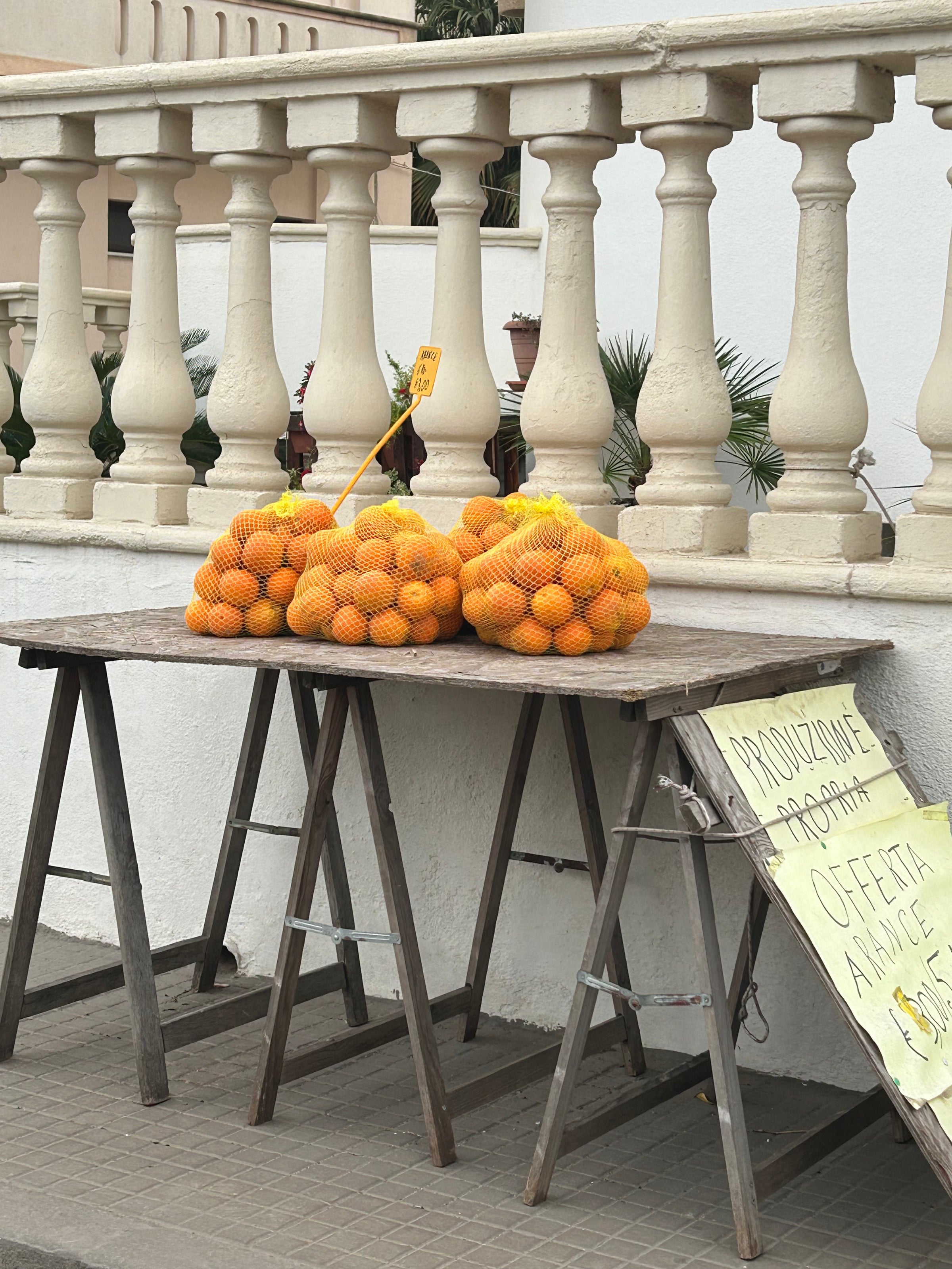 Bags of oranges are stacked on a wooden table in front of a white balustrade. A handwritten sign and a price tag are visible, suggesting the oranges are for sale outdoors.