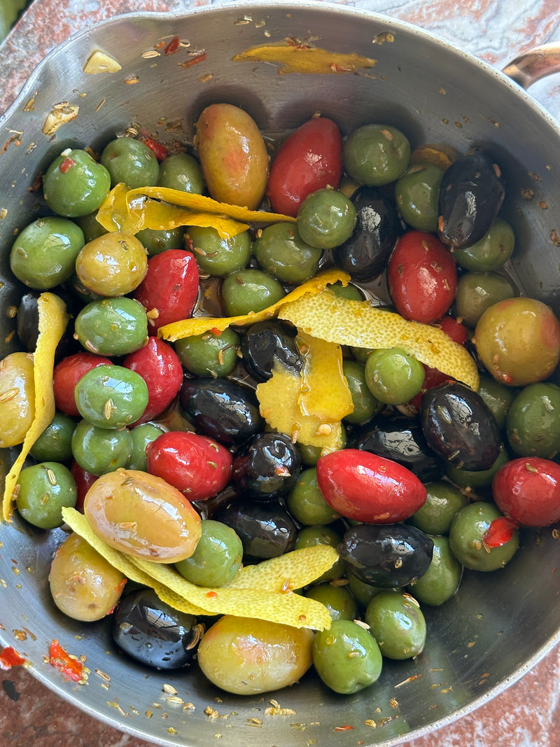 A metal bowl filled with a mix of green, red, and black olives, garnished with yellow citrus peels and small pieces of herbs and red chili. The olives appear glossy with oil.