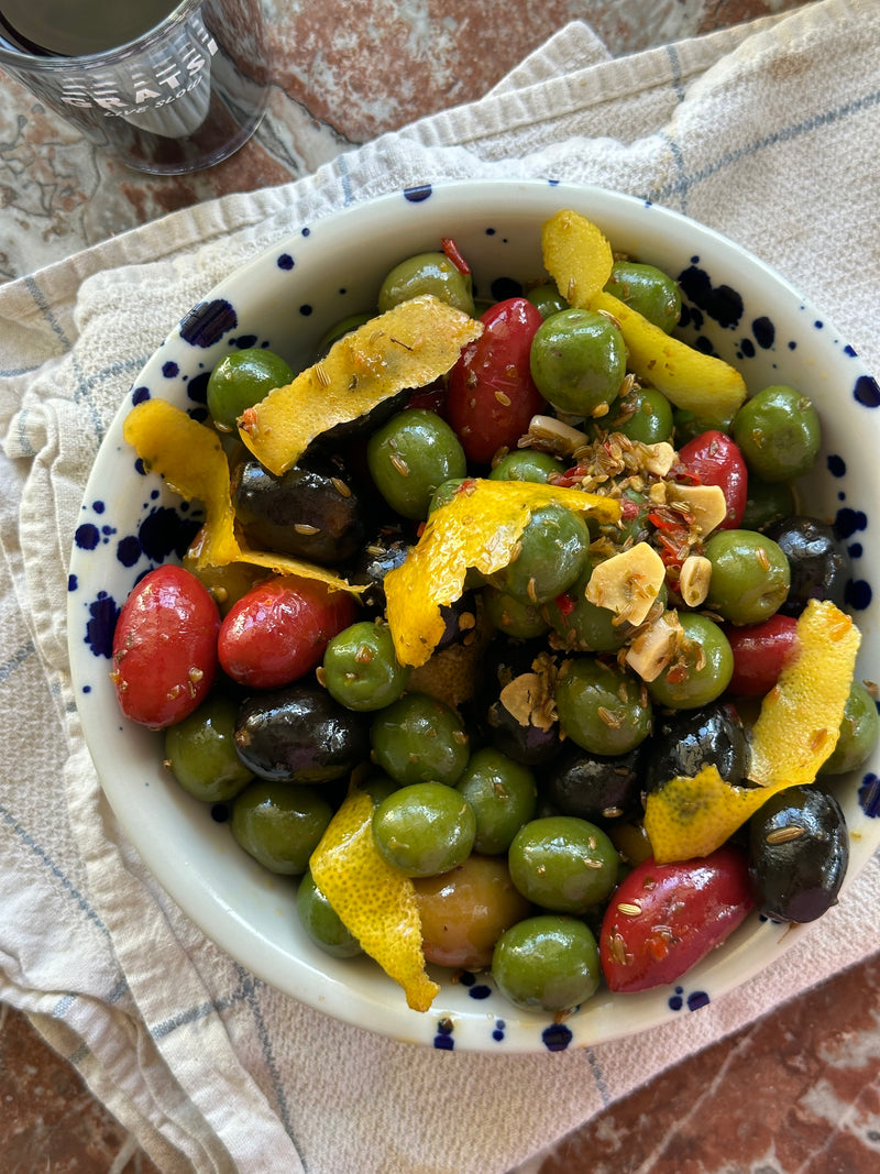 A bowl of mixed green, black, and red olives garnished with lemon peel and sliced garlic, served on a patterned dish towel on a marble surface.