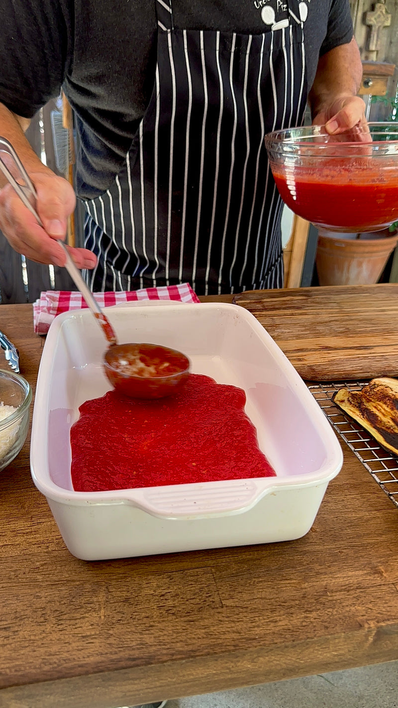 A person wearing a black-and-white striped apron ladles tomato sauce into a white rectangular baking dish on a wooden countertop. Other ingredients and utensils are visible nearby.