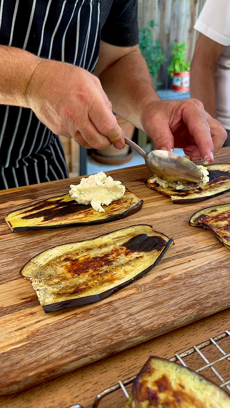 A person in a striped apron spoons creamy filling onto grilled eggplant slices placed on a wooden board, preparing a dish by hand. Another person stands nearby, assisting.