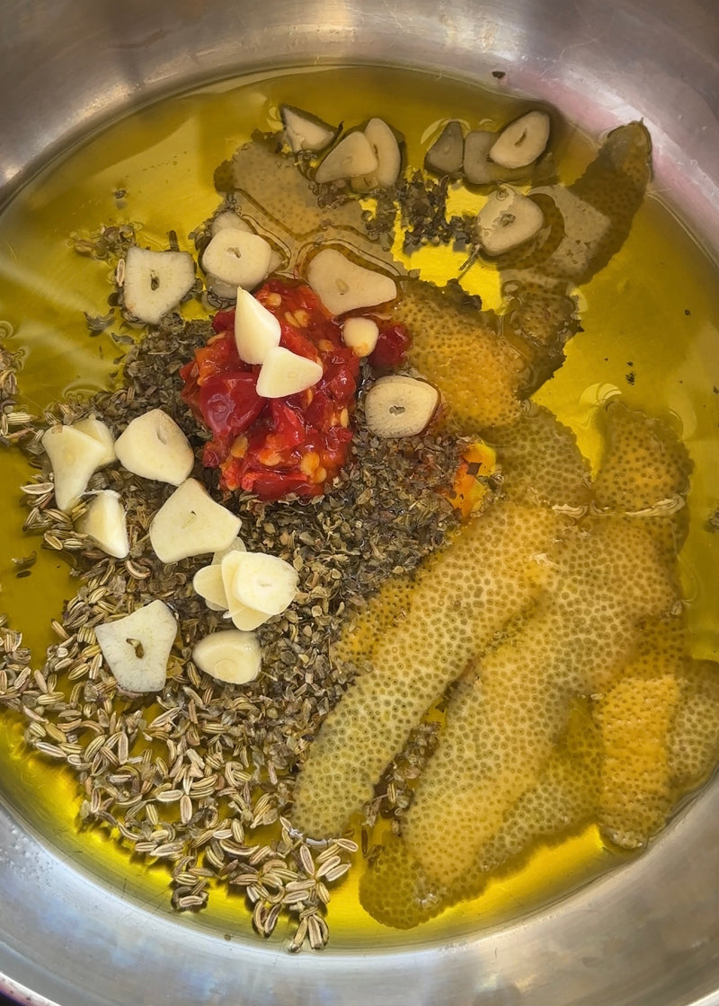 A metal pan with olive oil, fennel seeds, dried herbs, sliced garlic, red chili paste, and strips of lemon zest before cooking.