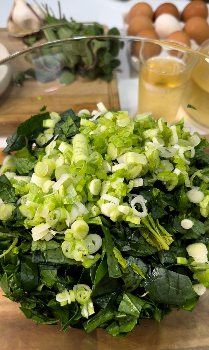 A bowl filled with chopped leafy greens and sliced green onions sits on a counter. In the background, eggs, a glass of light-colored liquid, and additional produce are visible.