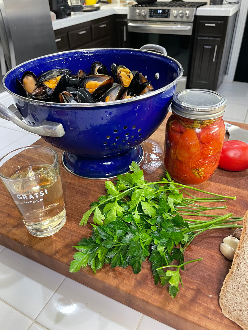 A blue colander filled with mussels on a wooden counter, next to a glass of white wine, a jar of whole peeled tomatoes, fresh parsley, garlic cloves, and a tomato, in a modern kitchen.