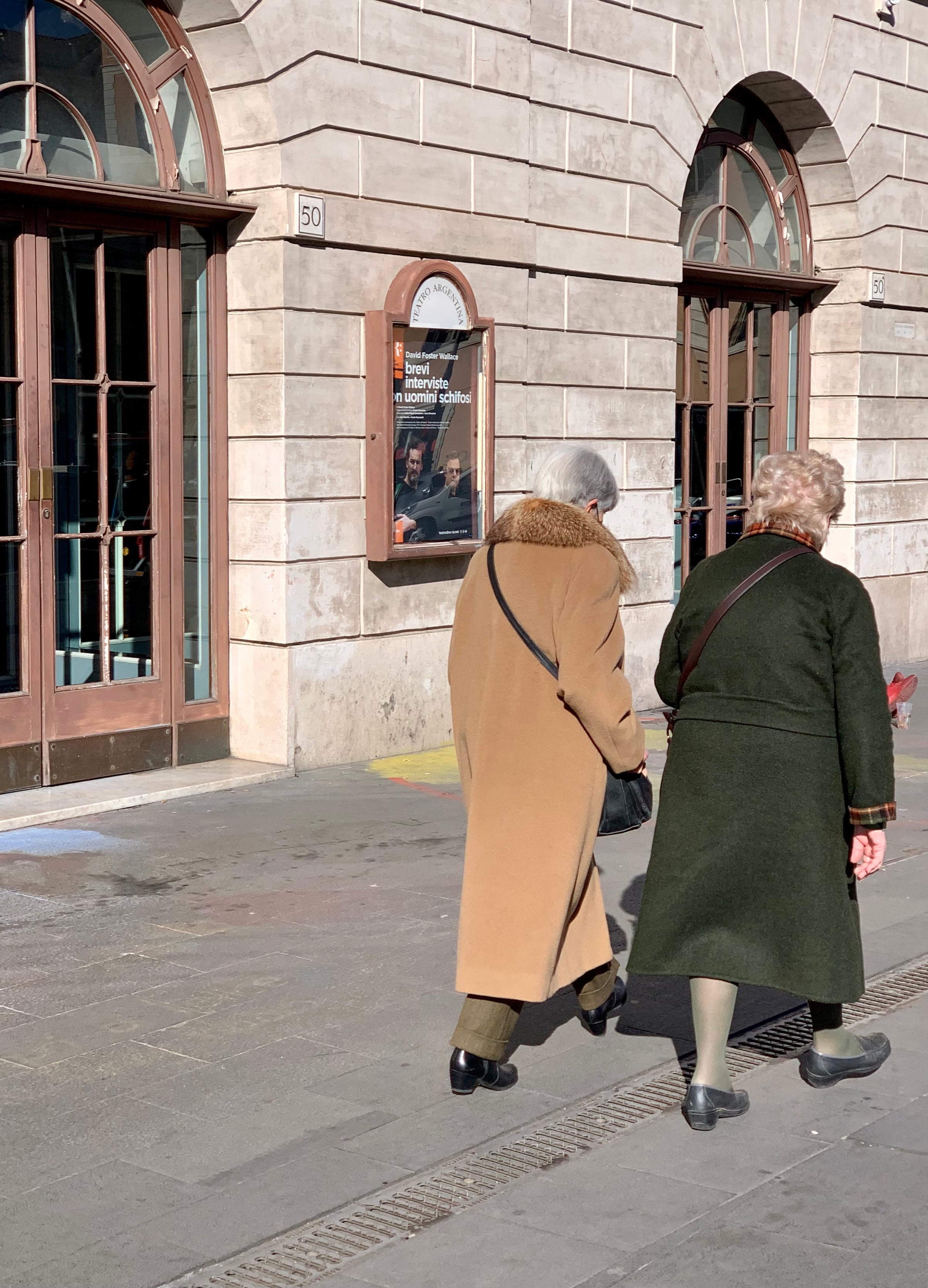 Two elderly people wearing long coats walk side by side on a sunny sidewalk next to a stone building with arched windows and a poster on the wall behind them.