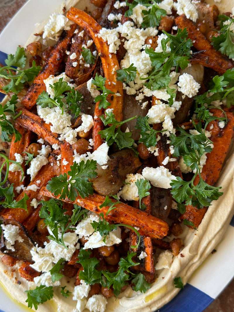 A close-up of a colorful dish with roasted carrots, chickpeas, and onions on a bed of hummus, topped with crumbled feta cheese and fresh parsley.