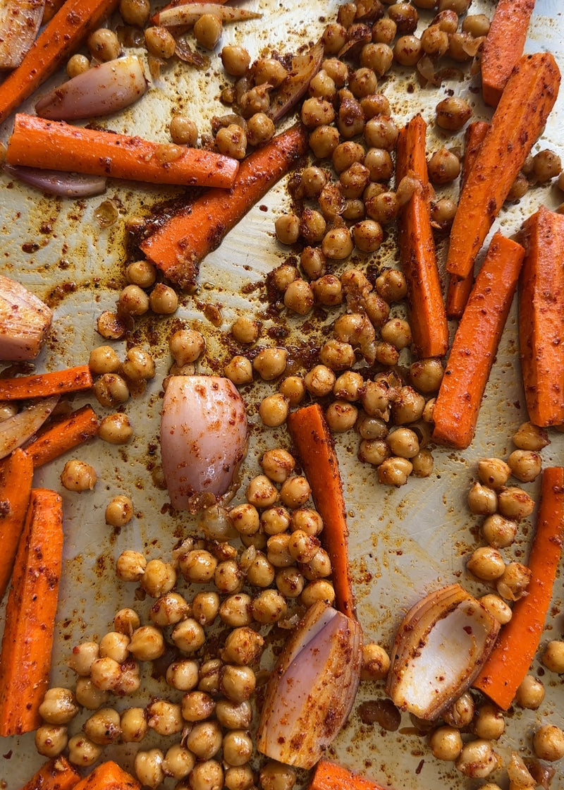 Chickpeas, carrot sticks, and shallot wedges seasoned with spices are spread out on a baking sheet, ready for roasting.
